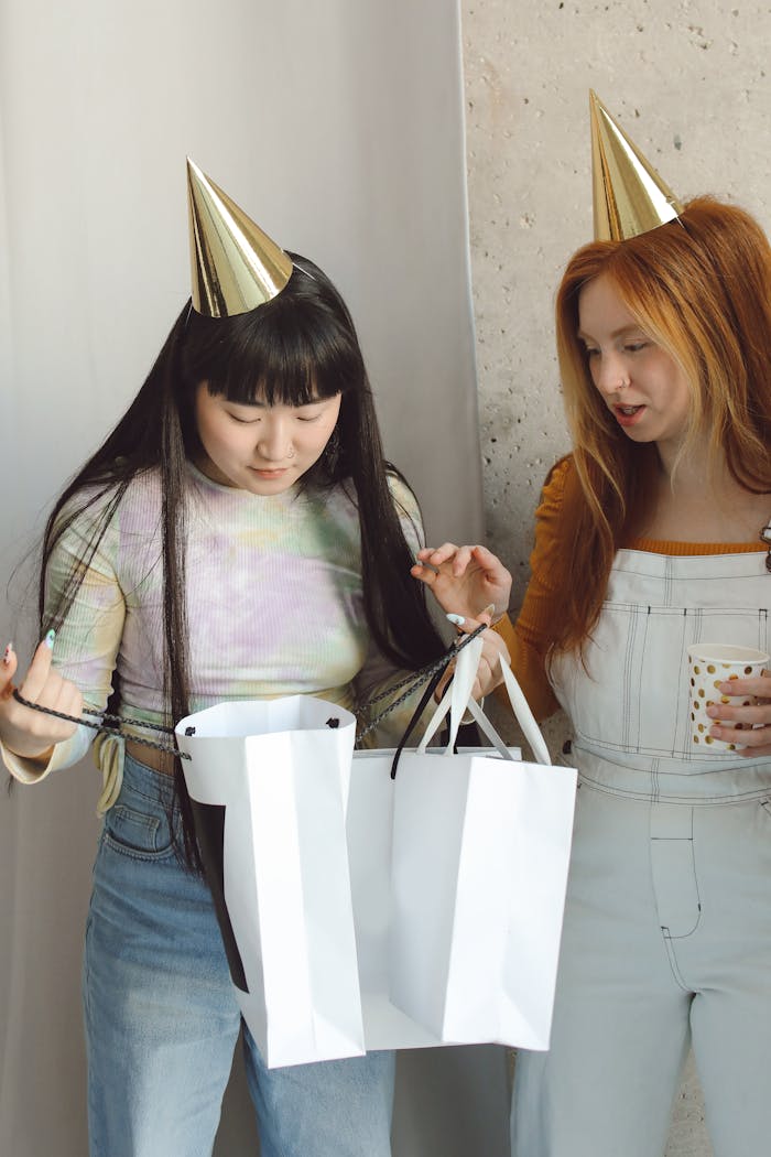 Two women wearing party hats exchange gift bags during a celebration indoors.